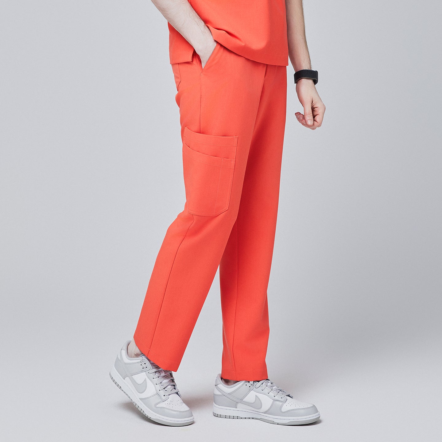 Angled side view of tiger orange scrub pants with spacious side pocket detailing and a tapered fit, paired with light gray sneakers, modeled against a neutral gray background,Tiger Orange