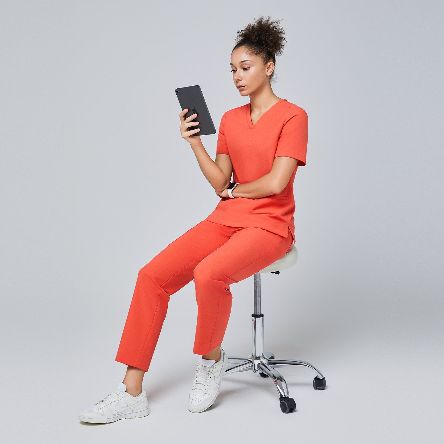 Woman in a tiger orange scrub set with straight-leg pants, seated on a white stool, holding a tablet, wearing white sneakers,Tiger Orange