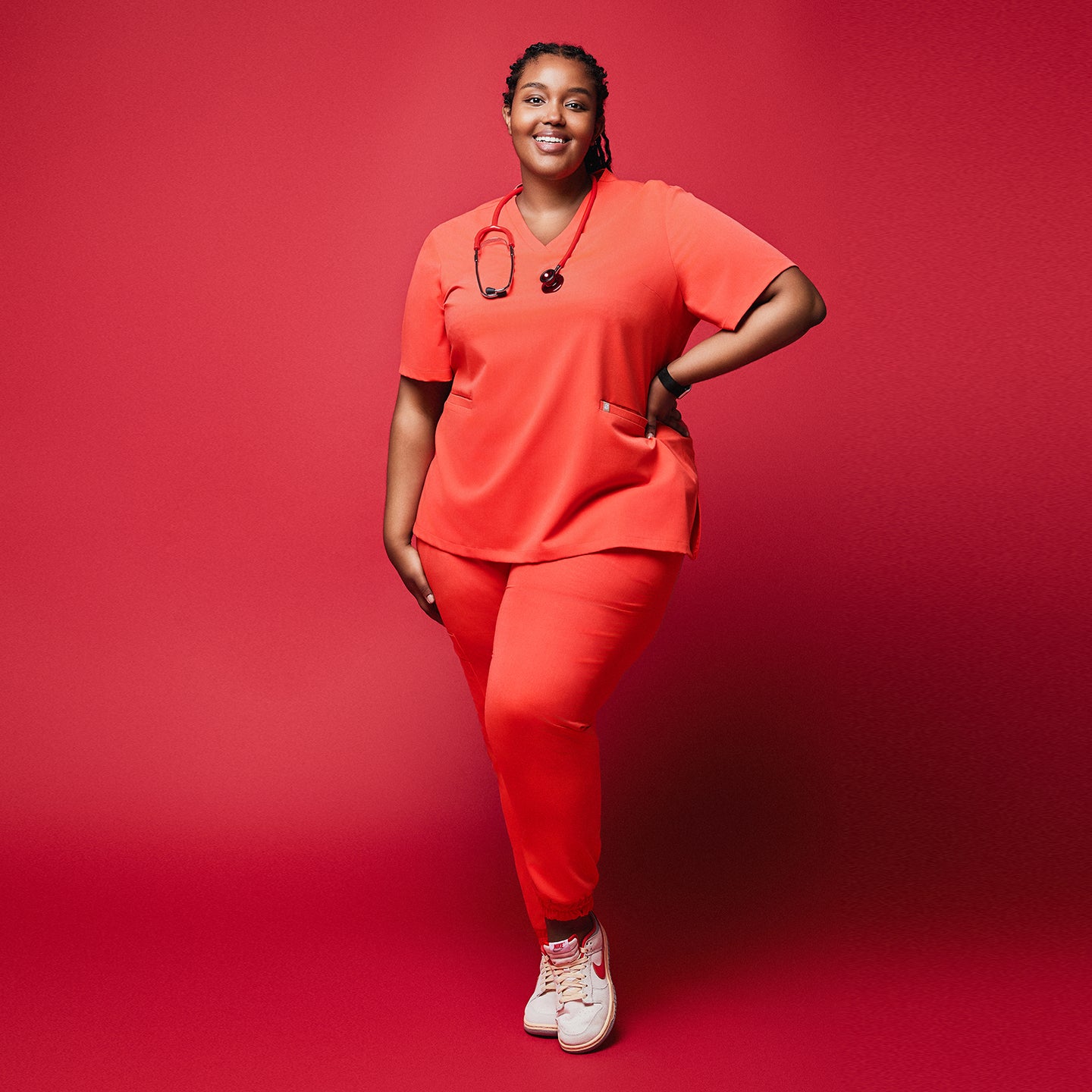 Woman wearing bright orange jogger scrubs with a stethoscope, smiling confidently against a red background,Tiger Orange