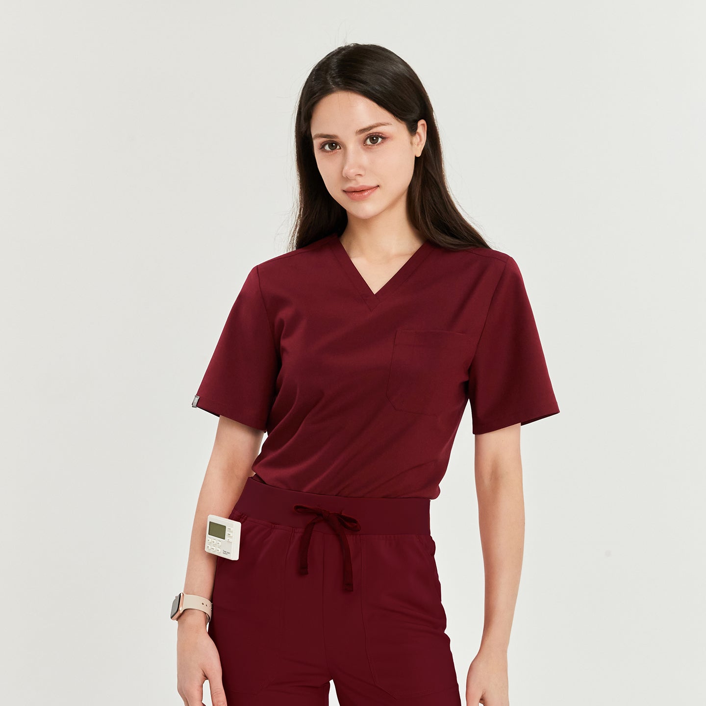 Woman in V-neck scrub top and pants with a medical device clipped to the waist, looking directly at the camera,Burgundy
