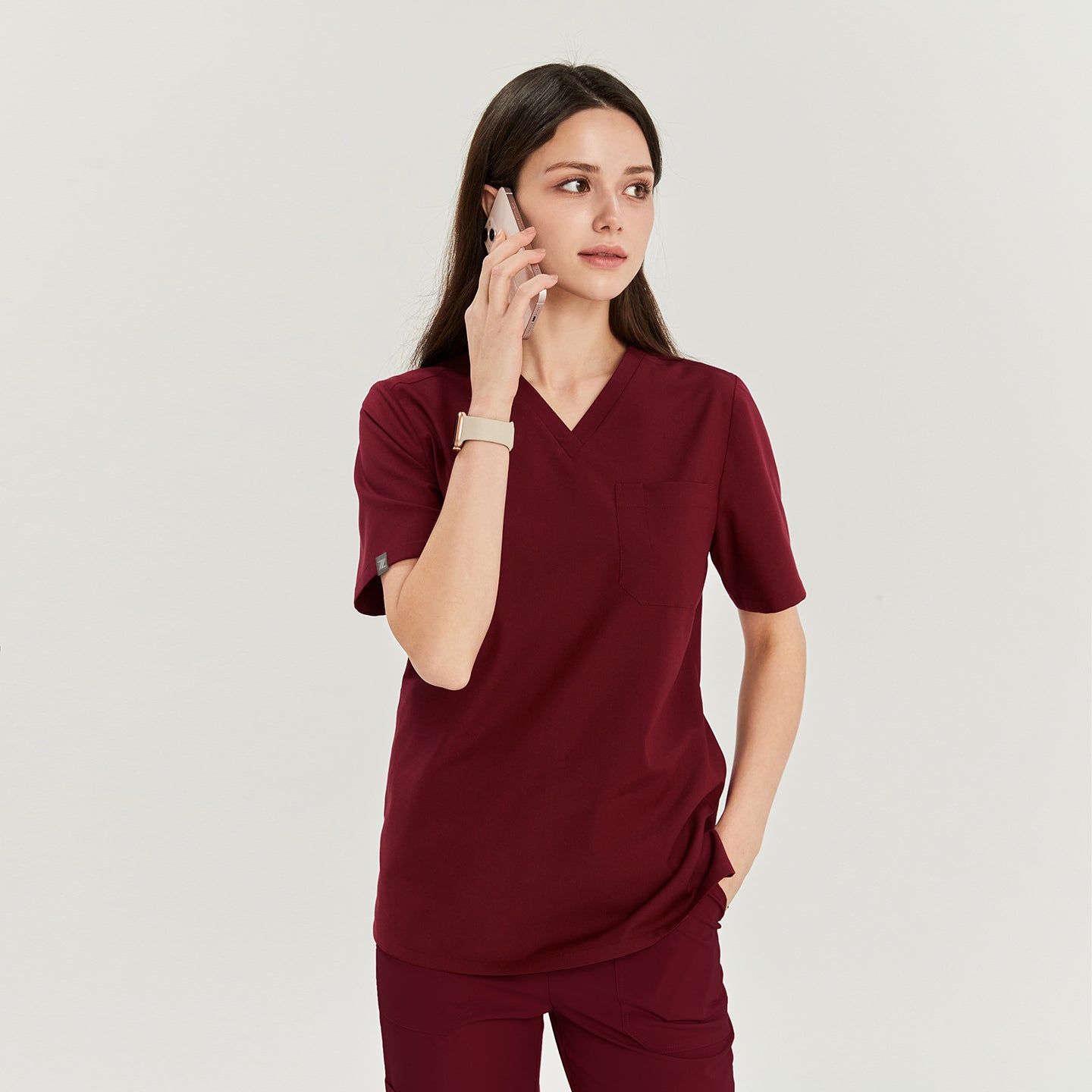  Woman in V-neck scrub top and pants with one hand in pocket, talking on a phone and looking to the side, Burgundy