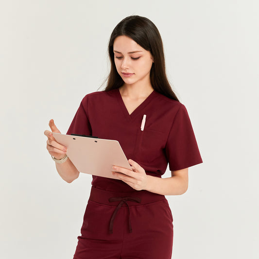 Woman in V-neck scrub top and pants with drawstring waist, holding a clipboard and pen, looking down,Burgundy