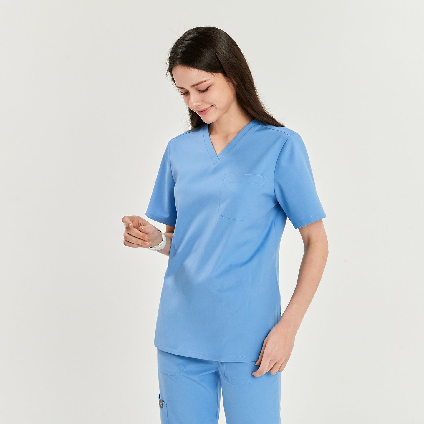 Woman wearing a V-neck scrub top and matching pants, looking down and smiling, with one hand on her side and the other hand slightly raised,Ceil Blue