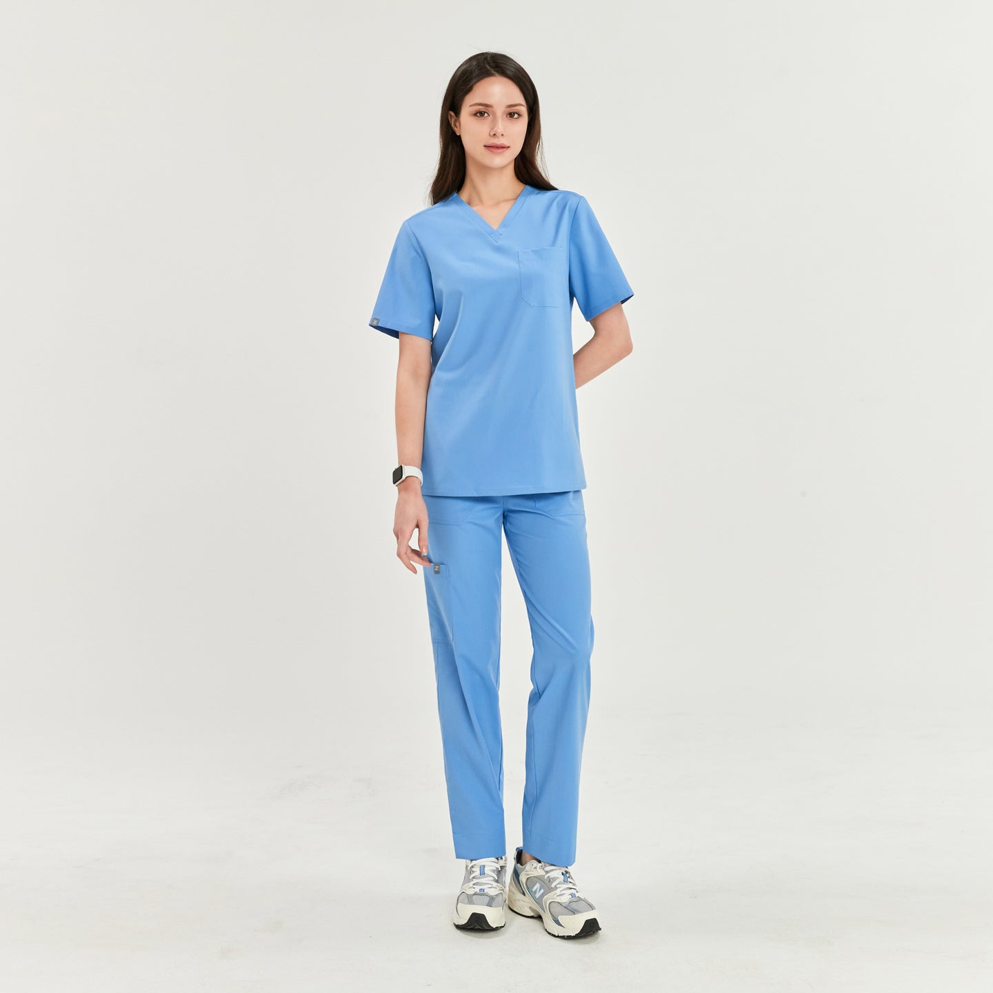 Woman standing in a V-neck scrub top and matching pants, hands at her sides, and looking straight ahead, Ceil Blue