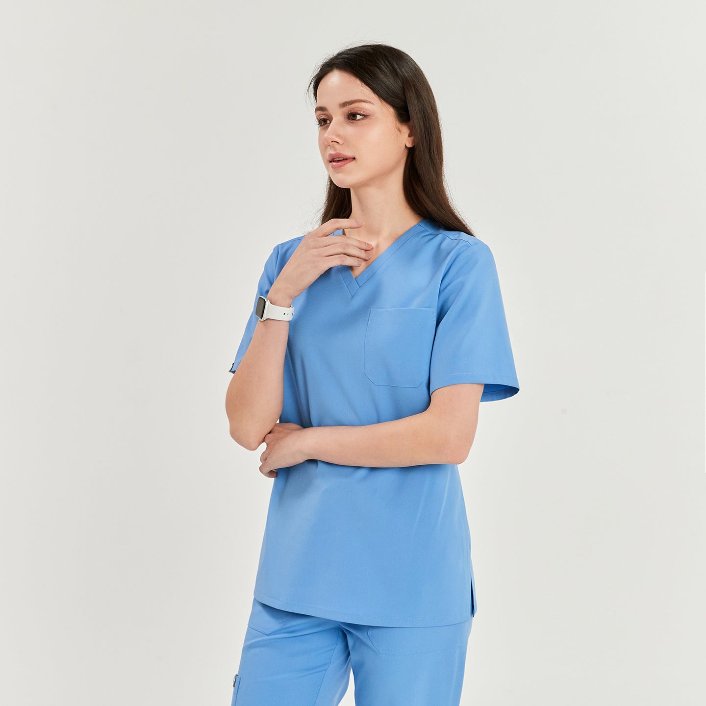  Woman wearing a V-neck scrub top and matching pants, standing with one hand touching her neck, looking to the side,Ceil Blue