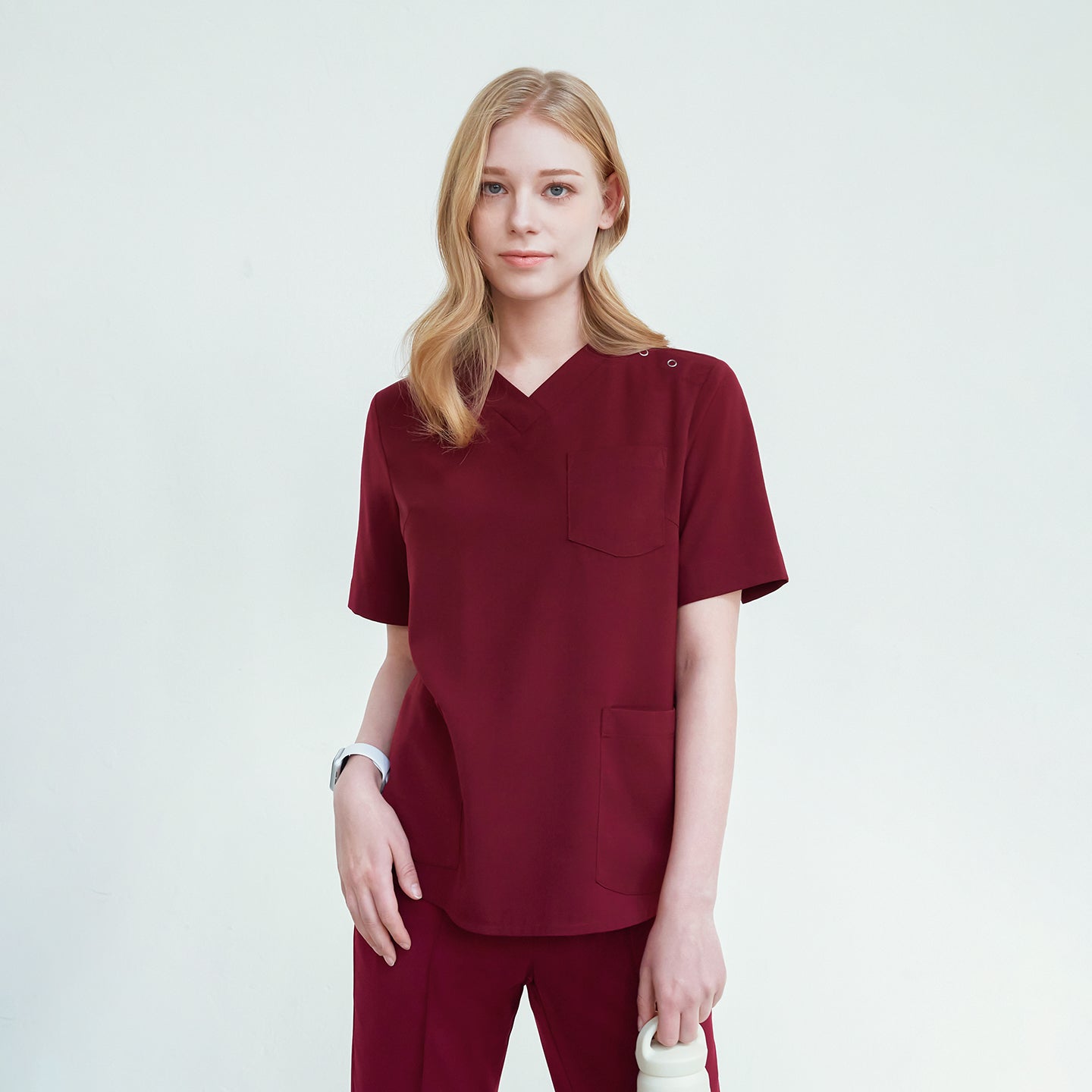 A woman wearing a scrub top with a V-neck and three buttons on the shoulder, paired with matching pants, standing against a plain background,Burgundy