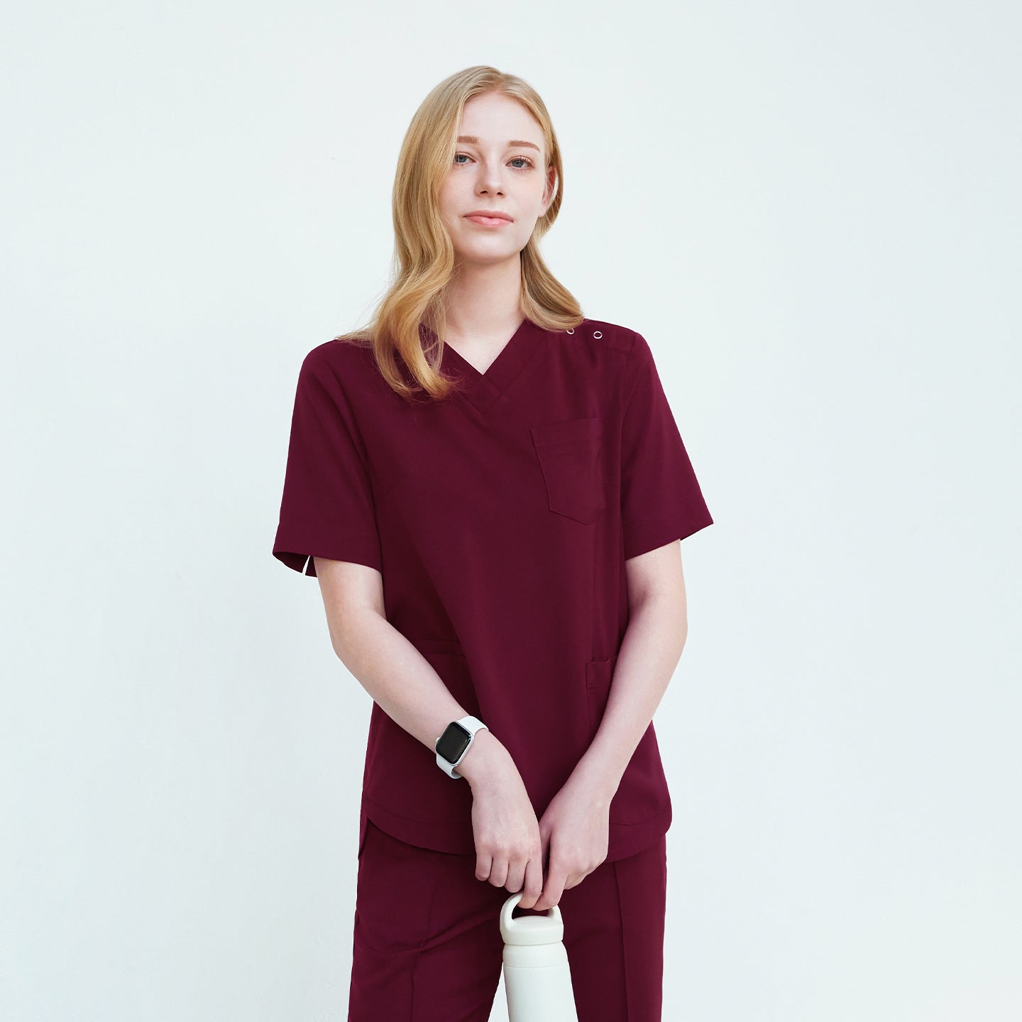 A woman in a V-neck scrub top with three buttons on the shoulder, holding a white water bottle and wearing a smart watch, standing against a plain background,Burgundy