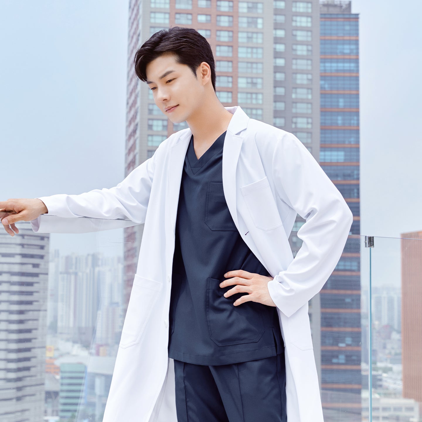 Man in a long lab coat over a front zipper scrub top with chest and side pockets, standing with one hand on a railing and the other on his hip, cityscape in the background,White