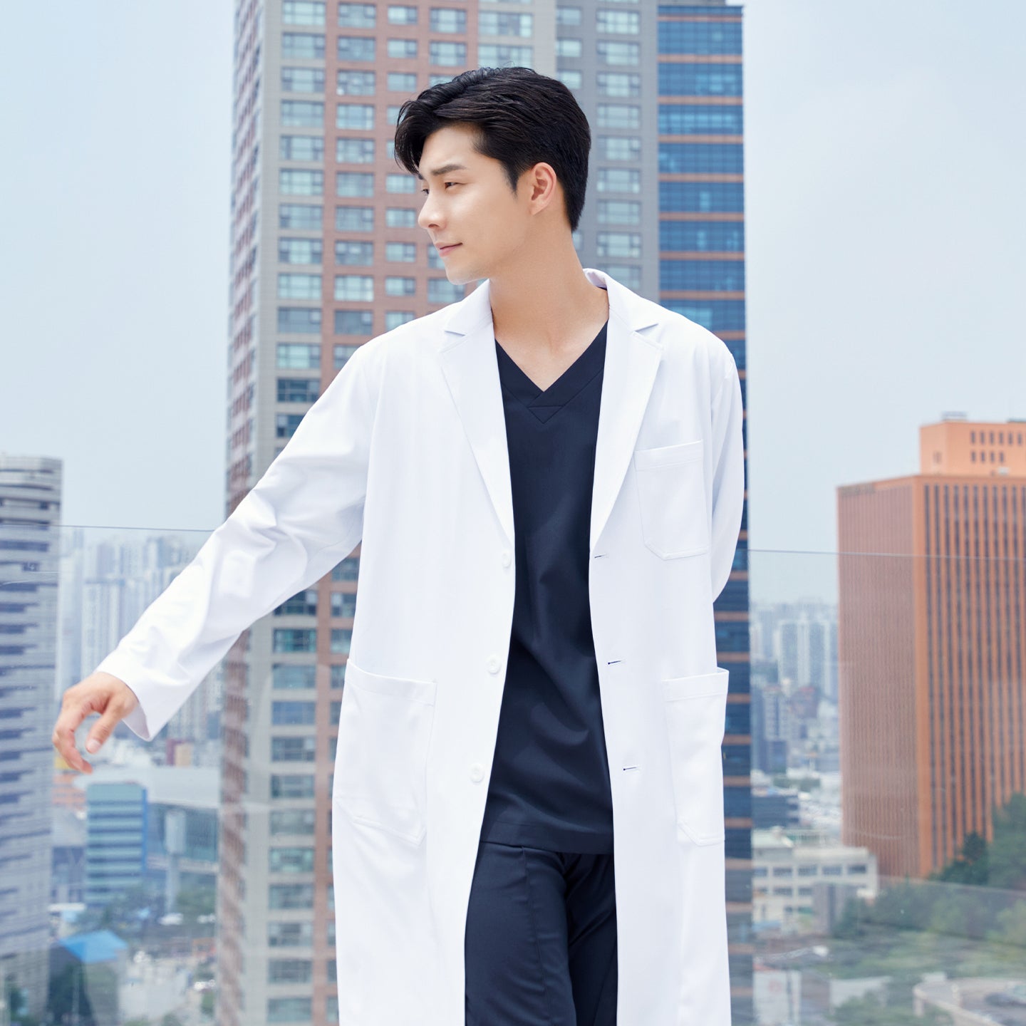 Man in a long lab coat over a front zipper scrub top with chest and side pockets, walking with a cityscape in the background,White