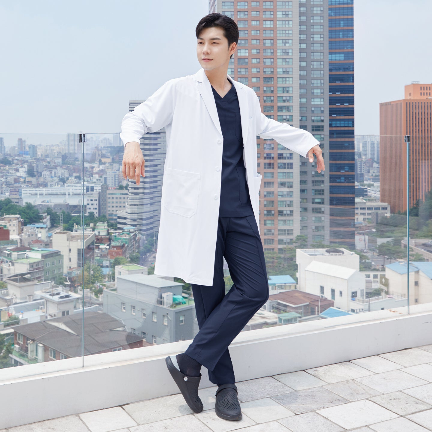 Man in a long lab coat over a front zipper scrub top with chest and side pockets, standing on a rooftop with a cityscape in the background,White