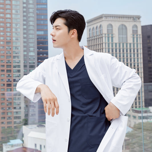 Man in a long lab coat over a front zipper scrub top with chest and side pockets, leaning on a railing with a cityscape in the background,White