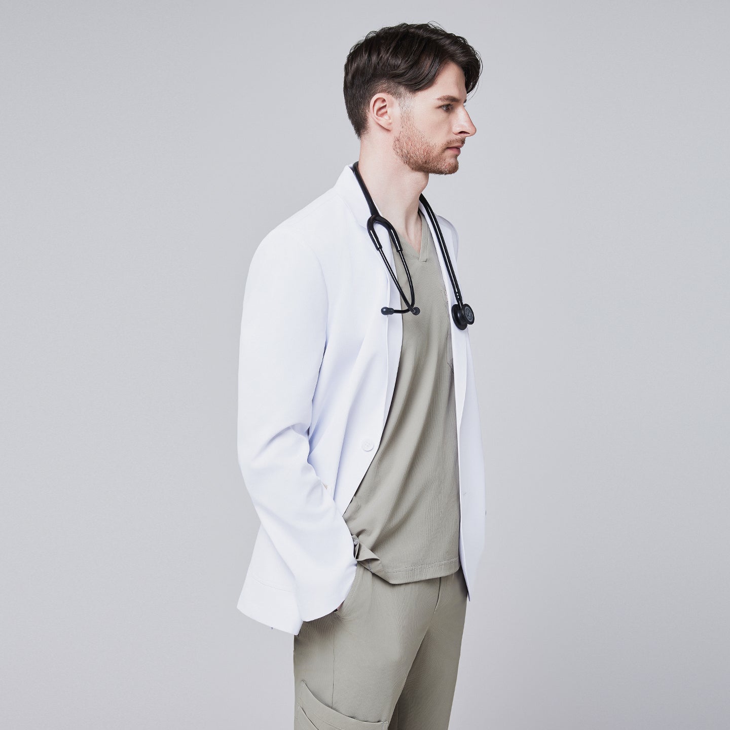 Male model in a white short lab coat over an olive green scrub set, posing in a profile view with hands in pockets,White