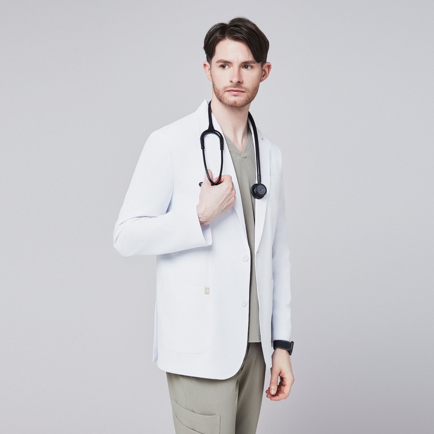 Male model in a white short lab coat over an olive green scrub set, holding a stethoscope with a thoughtful expression,White