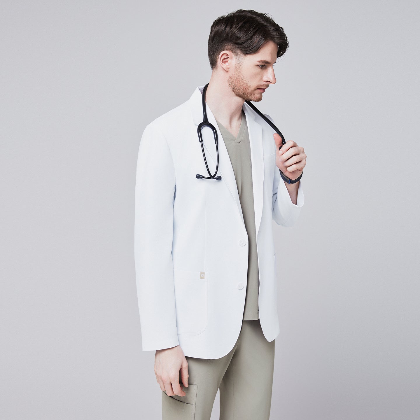 Male model wearing a white short lab coat over an olive green scrub set, holding a stethoscope in a side pose,White